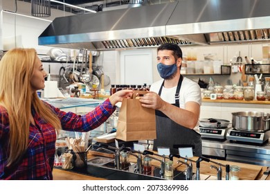 Owner Wearing Apron And Protective Face Mask Serving Takeaway Food To Customer At Counter In Small Family Eatery Restaurant During Covid 19 Restrictions. Selective Focus