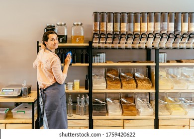 Owner Woman With Rag Cleaning Glass Dispenser With Metal Tap With Oil. Woman Assistant In Zero Waste Shop. Weighing Dry Goods In Plastic Free Grocery Store. Sustainable Shopping At Local Business.