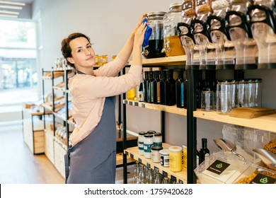 Owner Woman With Rag Cleaning Glass Dispenser With Metal Tap With Oil. Woman Assistant In Zero Waste Shop. Weighing Dry Goods In Plastic Free Grocery Store. Sustainable Shopping At Local Business.