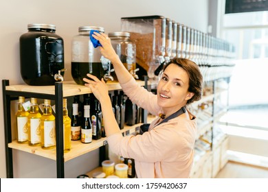 Owner Woman With Rag Cleaning Glass Dispenser With Metal Tap With Oil. Woman Assistant In Zero Waste Shop. Weighing Dry Goods In Plastic Free Grocery Store. Sustainable Shopping At Local Business.