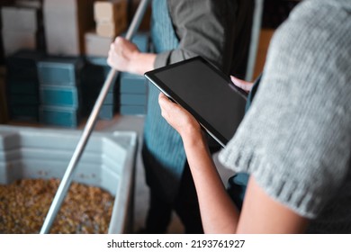 Owner working on a digital tablet in a winery while a wine maker press grapes to make wine. Woman worker doing research on the internet with technology in a manufacturing distillery warehouse. - Powered by Shutterstock