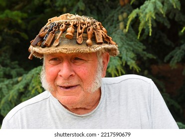 OXBOROUGH, NORFOLK, ENGLAND -  JULY  12, 2022: Older Man With Hat On Which Is Covered In Carved Wooden Love Spoons.