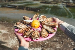 Oysters Plate, Cancale, Breton Capital Of Oysters, France. A Plate Of Fresh Oysters With Lemon Close-up In Female Hands On A Background Of An Oyster Farm In Cancale - Breton Capital Of Oysters, France