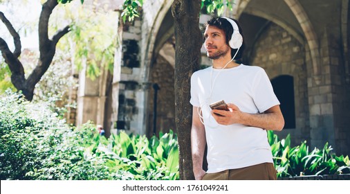 Pacified Young Male In Summer Outfit Listening To Audiobook In Headphones On Mobile Phone While Leaning On Tree In Park And Looking Away