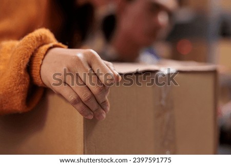 Package handler arm holding packed cardboard box in industrial warehouse. Storehouse employee picking order and preparing customer parcel for transportation with close up on worker hand