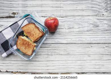 Packed Lunch Of Fresh Apple And Home Egg Salad Sandwich With Toasted Wheat Bread In A Glass Container. Healthy Vegetarian Diet Concept Over A Rustic White Wooden Background. Table Top View.