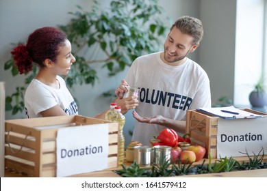 Packing Food For Needy. Young Bearded Man And Girl With Colored Hair, Standing At The Table With Donation Boxes, Holding A Can Of Canned Food, Smiling Volunteers.