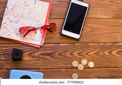 Packing Luggage For A Trip. A A Folded Map, A Mobile Phone, A Photo Camera, An Action Camera And Coins On A Rustic Wooden Background. Space For A Text Or Product Display, Top View.