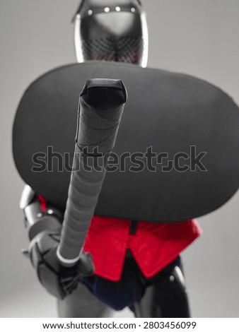 Padded sword and shield in close-up held by armored fighter in red jacket.