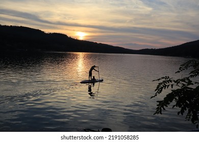 Paddle Board On Brno Lake In Evening Sundown. Sunshine Mirroring In Dark Water Of Dam On Svratka River During Evening After Hot Day. Forest On Embankment With Trees Is Black Before Night. Man And Kid.