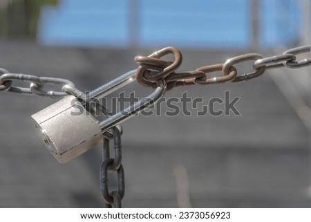 A padlock with a long hasp connecting a slightly rusty chain.The chain blocking the passage is secured with a padlock. Stairs blurry in the background .