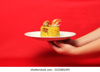 Paella rice is a traditional cuisine recipe from the city of Valencia Spain prepared with saffron, seafood, shrimp, vegetables served on a white plate on a red background qith hands serving - Powered by Shutterstock