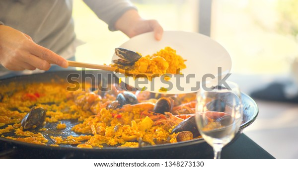 Paella\
traditional Spanish food. Person putts seafood paella from the fry\
pan to plate. Paella with with mussels, king prawns, langoustine\
and squids. Person cooking paella. Family\
Dinner