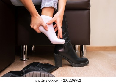 Pain In Feet From Uncomfortable Boots. A Woman Holds A Sore Foot With Hand Sitting On A Couch. Pain Caused By Wearing High-heeled Shoes. Discomfort From Walking In Heels For A Long Time. Orthodontics