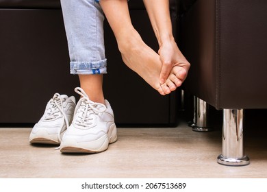 Pain In The Feet From Uncomfortable Shoes. A Woman Holds A Sore Foot With Hands Sitting On A Couch. Pain Caused By Wearing Sneakers. Discomfort From Walking For A Long Time. Orthodontics