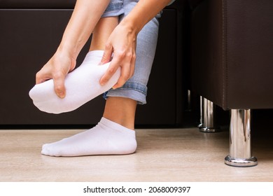 Pain In The Feet From Uncomfortable Shoes. A Woman Holds A Sore Foot In A White Sock With Hands Sitting On A Couch. Discomfort And Ache Caused By Walking For A Long Time. Orthodontics