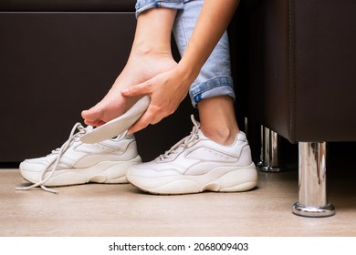 Pain in the feet from uncomfortable shoes. A woman i a white sneakers tries on a shoe insole sitting on a couch. Discomfort and ache caused by walking for a long time. Orthodontics