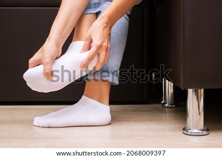 Pain in the feet from uncomfortable shoes. A woman holds a sore foot in a white sock with hands sitting on a couch. Discomfort and ache caused by walking for a long time. Orthodontics