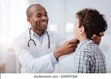 Pain in throat. Handsome afro American male doctor looking at boy while smiling and checking his throat