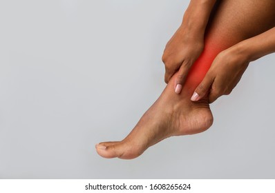 Pain in woman ankle. Black girl massaging her leg to stop ankle-ache, grey background, copy space