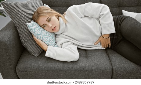 A pained blonde woman with blue eyes dressed in a white sweater resting on a couch indoors, holding her stomach. - Powered by Shutterstock