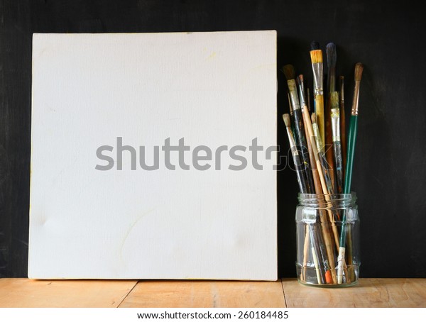 paint brushes in jar and blank canvas over\
blackboard background\
