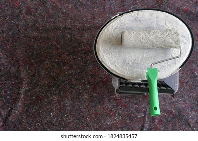 A Paint Roller Soaked With White Color And Put On The Lid Of A Paint Container With Lot Of Copy Space. The Equipment Is Placed On A Protective Carpet To Prevent Damages And Color Spots On The Floor. 