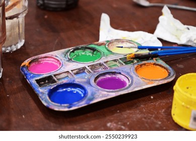 A paint table and brushes display an array of colorful watercolor paints, their lids open and ready for the artist's next work