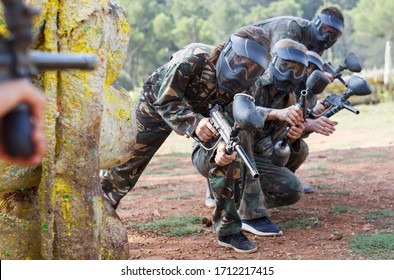 Paintball players of one team in camouflages and masks aiming with gun in shootout on battlefield