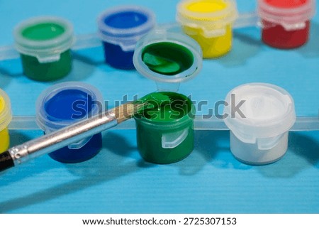 a paintbrush dipping into a small open pot of green paint, surrounded by other colorful plastic containers on a blue surface.