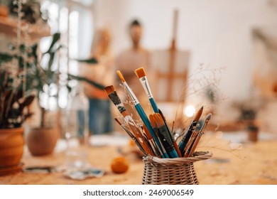 Paintbrushes in container at creative atelier with artistic couple painting on easel in a blurry background. Selective focus on various paintbrushes at creative art studio with painters painting. - Powered by Shutterstock