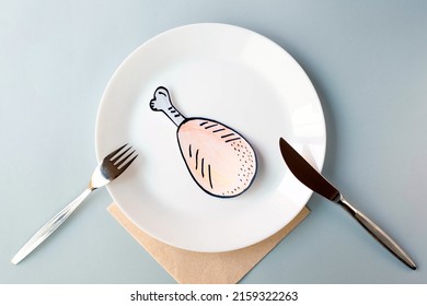 A painted chicken leg lies on a white plate served with a table knife and fork on a gray background. The topic of economic sanctions and food shortages. View from the top point.