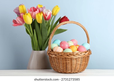 Painted Easter eggs in wicker basket on white wooden table and beautiful tulips in vase against light blue background