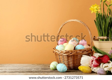 Painted Easter eggs in wicker basket and beautiful flowers on wooden table against pale brown background, closeup. Space for text