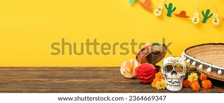 Painted human skull for Mexico's Day of the Dead (El Dia de Muertos), sombreros and flowers on table against yellow background with space for text