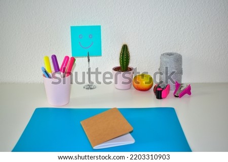 A painted smiley face on a blue piece of paper stands on the desk next to a notebook, a small cactus, an apple and markers