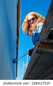 Painter Adult Mature Redhead Woman With Curly Red Hair And Sunglasses, Stands On A Facade Scaffolding And Paints Blue Color Paint On The House Wall Painting With Painter Roller Under The Blue Sky