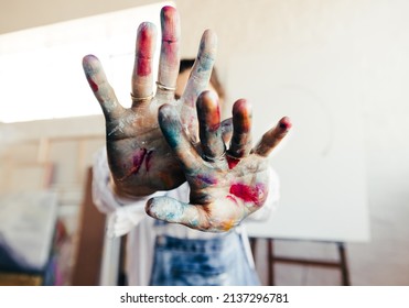 Painter blocking the camera from her face with her colour painted hands. Unrecognizable female artist standing in her art studio with her blank canvas in the background.
