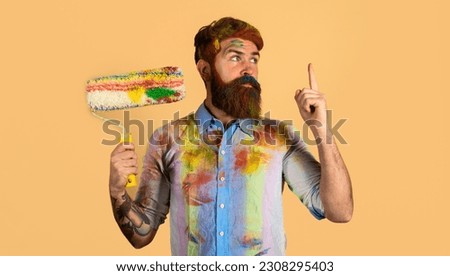 Painter man with paint roller pointing finger up. Professional painter, decorator, builder worker with paintroller. Handsome bearded worker, repairman or handyman in dirty dye shirt with paint roller.