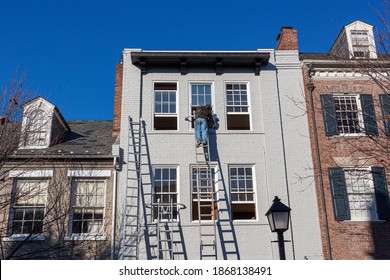 A Painter Is On Top Of An Industrial Size Aluminum Extension Ladder Painting The Exterior Surface Of An Old Brick House. He Wears No Safety Equipment While Working At This Dangerous Height.