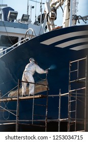 painter painting the hull of a ship , the painter is on a scaffold and is painting with a mask on his face and a white coveral or worker painting a ship on a scaffold