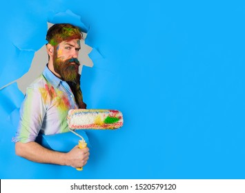 Painter. Painter with painting roller through hole in paper. Handsome bearded worker with paint roller. Professional painter in dirty shirt making hole in paper wall. Serious painter man. Copy space.