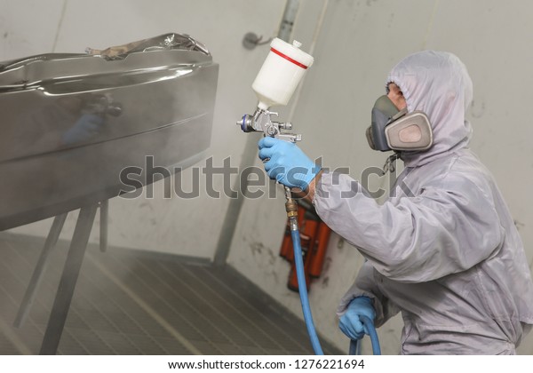 the
painter paints the car detail in the painting chamber. the paint is
spraying from the spray gun on the car part. car service worker in
mask and overalls is painting a detail of the
car