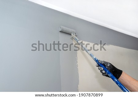 The painter paints the room gray with a paint roller on an extended stick at home.
