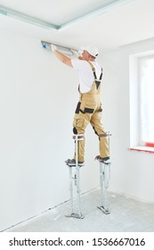 Painter In Stilts With Putty Knife. Plasterer Smoothing Ceiling Surface At Home Renewal