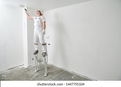 Painter worker on stilts with roller painting ceiling into white