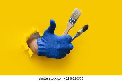 The painter's hand in a blue knitted cloth glove holds two bristle brushes and shows a big stick up (like). Ripped hole in yellow paper, copy space. The concept of a worker, a labor migrant, a master. - Powered by Shutterstock