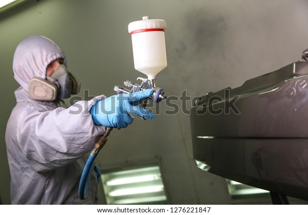 the painter\'s hand paints the car detail in the\
painting chamber. the paint is spraying from the spray gun on the\
car part. car service worker in mask and overalls is painting a\
detail of the car