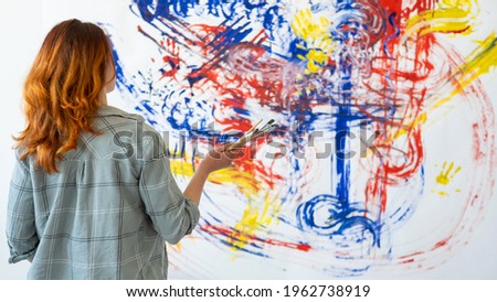 Painting art. Interior decor. Creative design. Back view portrait of inspired redhead woman painter with brushes looking at blur colorful blue red yellow abstract artwork on white wall background.
