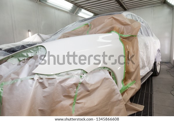 Painting the car body parts of\
the driver\'s white fender in the spray booth in the body repair\
workshop, other elements are covered with film and paper from\
splashing.